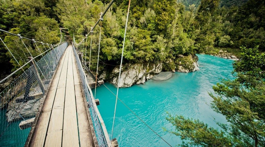 Wooden plank and steel susupension bridge above a river with turquoise water flowing between lush green valley on rocks